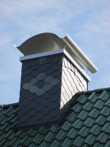 Schornsteinkopf Schornstein auf einem Dach mit grünen Schindeln und einem weißen Kaminaufsatz.