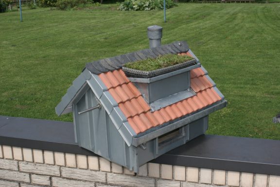 Vogelhaus Ein kleines graues Haus mit einem roten Ziegeldach und bepflanztem Schornstein.