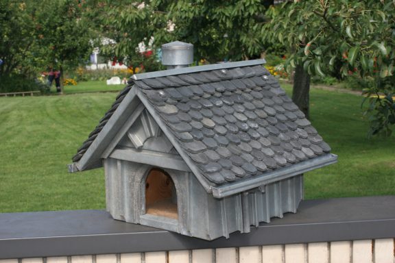 Vogelhaus Ein Holzhäuschen mit schiefergedecktem Dach auf einer Mauer, umgeben von Grün.