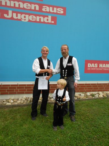 Lennart Recker Lossprechung Zwei Männer in traditioneller Kleidung schütteln sich die Hände, ein Junge steht dazwischen.