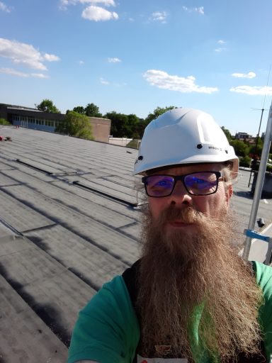 Titus Recker Ein Mann mit Brille und Helm steht auf einem Dach unter blauem Himmel.