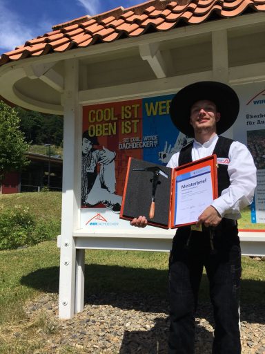 Lennart Recker Meisterprüfung Mann in traditioneller Kleidung hält eine Urkunde vor einem Plakat im Freien.